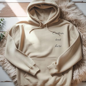 May include: A beige hooded sweatshirt featuring long sleeves, a front kangaroo pocket, and matching drawstrings. The chest area displays the black printed text "Custom text here" in a cursive font. The garment is constructed with ribbed cuffs.