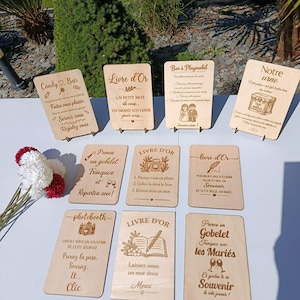 Peut inclure: Un ensemble de dix panneaux rectangulaires en bois clair avec des textes et illustrations gravés en français pour des mariages, incluant des indications pour candy bar, livre d'or, photobooth et urne, présentés sur des petits supports.