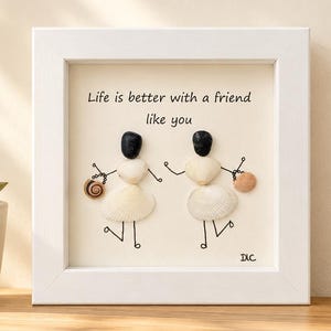 Peut inclure: Un cadre carré blanc contenant une œuvre d'art en galets représentant deux personnages faits de coquillages et de fil de fer noir. Les personnages ont les bras levés et les jambes en mouvement. Le texte "Life is better with a friend like you" est imprimé.