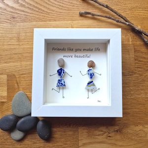 May include: A white framed artwork with two figures crafted from stones and ceramic fragments. The artwork includes the text "Friends like you make life more beautiful". The figures represent friends. The background is a light color, and the frame is square.