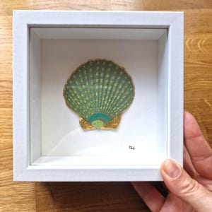 Può includere: Un'opera d'arte incorniciata con una conchiglia verde e oro. La conchiglia è esposta in una cornice bianca. La conchiglia ha una forma a ventaglio con dettagli intricati e un bordo dorato. L'opera d'arte è su uno sfondo bianco.