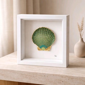 Könnte beinhalten: Ein gerahmtes, dekoratives Muschelgehäuse. Die Muschel ist grün mit goldenen Akzenten und einem türkisfarbenen fächerförmigen Detail. Es ist in einem weißen quadratischen Rahmen eingefasst. Das Kunstwerk wird auf einer hellen Oberfläche präsentiert.