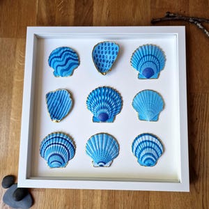 Op de afbeelding: Een witgelijst kunstwerk met negen blauwe schelpen met gouden rand. De schelpen hebben verschillende patronen en blauwtinten. Het kunstwerk wordt op een witte achtergrond gepresenteerd.