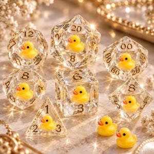 May include: A set of seven clear polyhedral dice, each containing a miniature yellow rubber duck inside. The dice feature gold-coloured numbers, including a d20, d12, d10, d8, d6, and d4. Two loose miniature yellow rubber ducks are placed nearby.