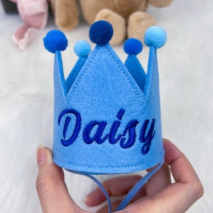 May include: A light blue felt crown embroidered with the name "Daisy" in a darker blue script. The crown has five peaks, each with a blue pom-pom. A blue ribbon is attached for securing the crown.
