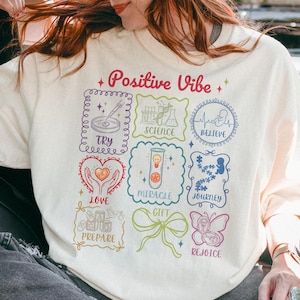Op de afbeelding: Crèmekleurige sweatshirt met de woorden "Positive Vibe" in roze en diverse kleurrijke grafische ontwerpen. De ontwerpen omvatten wetenschappelijke illustraties, harten en een vlinder, met woorden als "Try", "Love" en "Believe".