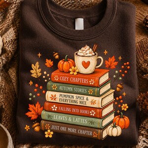 May include: A dark brown crewneck sweatshirt featuring a graphic of a stack of six books topped with a mug of whipped cream. The book spines are printed with the text Cozy Chapters, Autumn Stories, Pumpkin Spice & Everything Nice, Falling Into Books, Leaves & Lattes, and Just One More Chapter, surrounded by autumn leaves, pumpkins, and acorns.