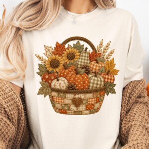 以下が含まれることがあります： クリーム色のTシャツで、中央に編みかごに入った様々なカボチャ、ヒマワリ、秋の葉、小麦の茎が描かれています。かごはパッチワーク柄で、前面に小さなハートのディテールがあしらわれています。