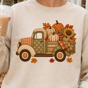 May include: A cream-colored crewneck sweatshirt featuring a graphic of a vintage pickup truck with a patchwork design. The truck bed is filled with pumpkins, a sunflower, and autumn leaves, with additional fall leaves scattered below the truck.