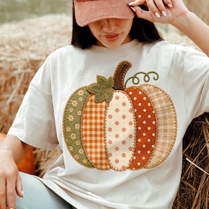 Op de afbeelding: Een crèmekleurig T-shirt met korte mouwen en een grote pompoenafbeelding in het midden. De pompoen bestaat uit vijf verticale segmenten, elk met een ander patroon, waaronder groene bloemen, oranje ruiten, crèmekleurige bloemen, oranje stippen en lichtbruine ruiten.