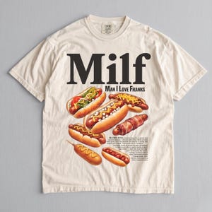 Pode incluir: Camiseta creme com a palavra "Milf" em letras pretas grandes e "Man I Love Franks" abaixo. A camisa exibe ilustrações de vários cachorros-quentes com coberturas, incluindo mostarda, picles e bacon.