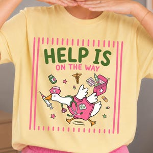 Può includere: Una t-shirt giallo pallido con la grafica di un'oca bianca vestita da infermiere con una casacca e un cappellino rosa e uno stetoscopio. Il testo HELP IS ON THE WAY è circondato da icone mediche come una siringa e un kit di pronto soccorso.