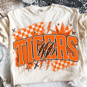 Puede incluir: Una camiseta de manga corta color crema con un gran gráfico naranja que muestra la palabra TIGERS en letras de bloque negritas. El diseño incluye un patrón de tablero de ajedrez desgastado, rayos, estrellas y tres marcas de garras sobre el texto.