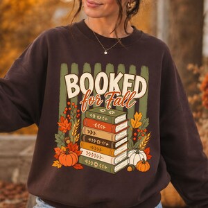 以下が含まれることがあります： ダークブラウンのクルーネックトレーナー。中央には「BOOKED for Fall」の文字と5冊の本が積み重なったグラフィックがプリントされています。デザインには、オレンジ色のカボチャ、どんぐり、麦の穂、色鮮やかな秋の葉といった秋をテーマにしたイラストが描かれています。