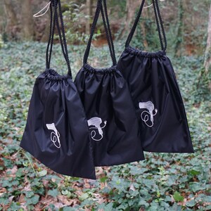 May include: Three black drawstring bags hanging from a red wooden bar. Each bag has a white embroidered design of a hairdryer with a cord.