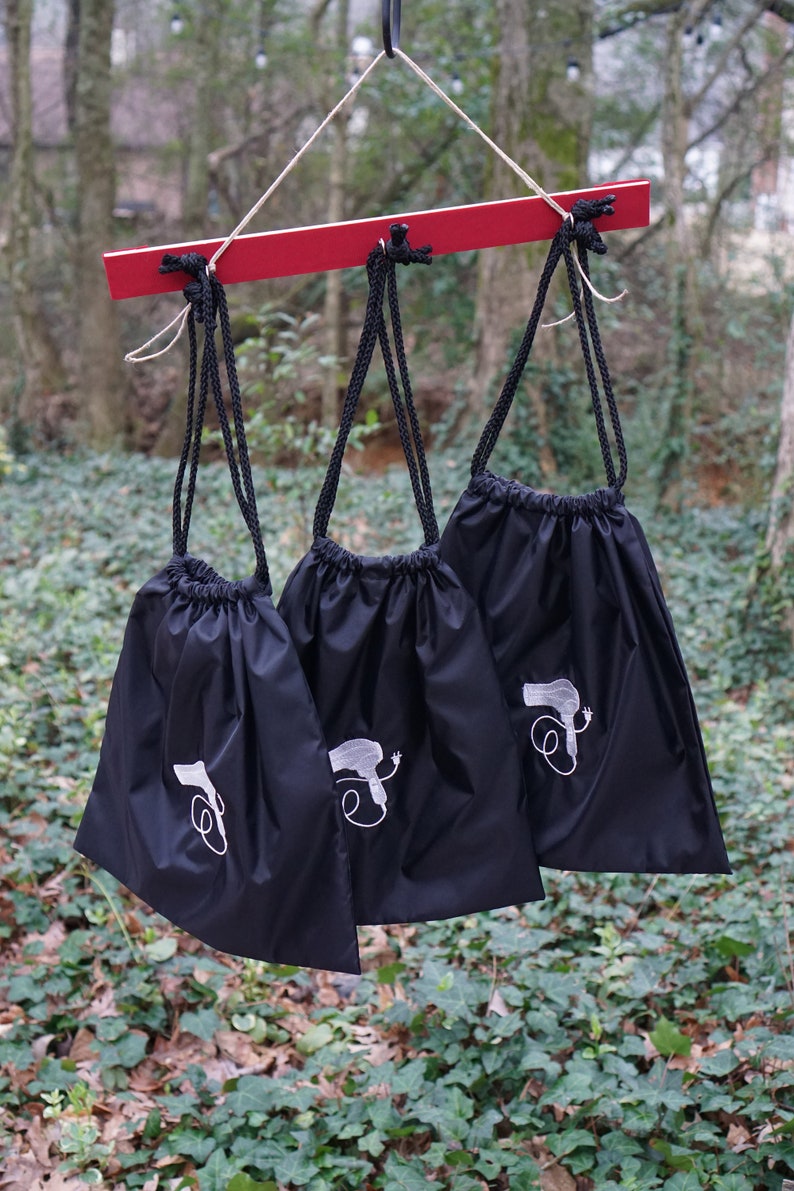 May include: Three black drawstring bags hanging from a red wooden bar. Each bag has a white embroidered design of a hairdryer with a cord.