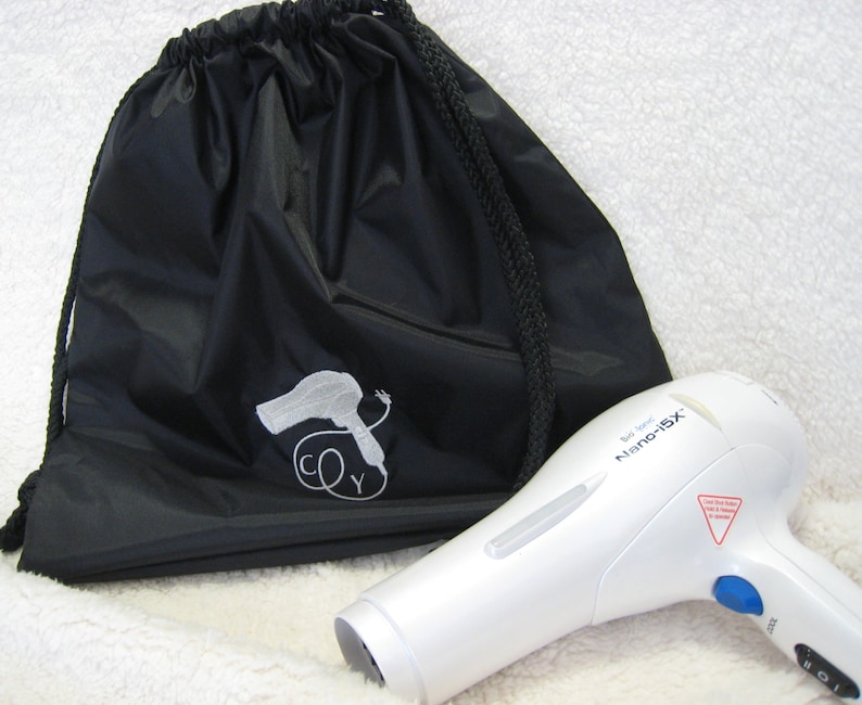 May include: A white hair dryer with the text "Tour Ionic Nano-iSX" on the side. The hair dryer is next to a black drawstring bag with a white embroidered image of a hair dryer and the letters "CY".