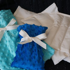 May include: Three heat packs, one blue, one turquoise, and one white, are tied with white ribbon bows. The heat packs are made of soft, plush fabric.
