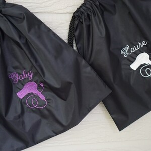 May include: Two black drawstring bags with a hairdryer graphic and personalized names. The bag on the left has the name "Gaby" embroidered in purple and the bag on the right has the name "Laure" embroidered in white.