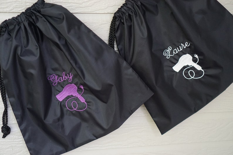 May include: Two black drawstring bags with a hairdryer graphic and personalized names. The bag on the left has the name "Gaby" embroidered in purple and the bag on the right has the name "Laure" embroidered in white.