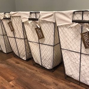 May include: Four wire laundry baskets with white fabric liners and brown labels. The labels read "Colors", "Darks", "Lights", and "Towels".