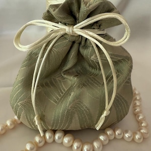 May include: A green fabric drawstring bag with a white satin ribbon tied in a bow. The bag is sitting on a white surface with a strand of white pearls.