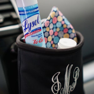 May include: A black car organizer with the initials "J.M.P." embroidered on the front. The organizer contains a blue and white patterned face mask, a bottle of Lysol Disinfectant Spray To Go, and a small white bottle.