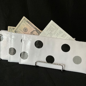May include: Three white zippered pouches with black and gray polka dots. The pouches are folded and stacked on top of each other. A metal handle is attached to the bottom pouch. The pouches are filled with cash and a receipt.