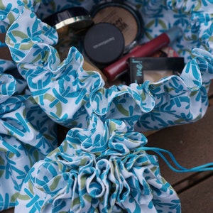 May include: A blue and white drawstring bag with a floral pattern. The bag is open and filled with makeup products, including a compact, a lipstick, and a powder brush.