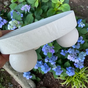 May include: White wooden pedestal with three rounded legs. The pedestal is sitting on a bed of blue flowers.