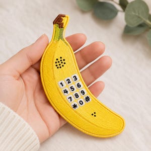 May include: A yellow banana-shaped patch with a phone keypad design. The patch features a brown stem, a speaker, and a keypad with numbers and symbols. The patch is a fun, quirky accessory.