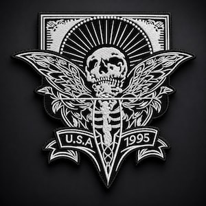 Può includere: Una toppa ricamata in bianco e nero raffigurante un teschio centrale con ali, una gabbia toracica e un design a raggiera. Un banner nella parte inferiore mostra il testo U.S.A e 1995. Il design è racchiuso in una cornice rettangolare con angoli decorati.