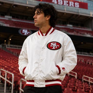 May include: A white satin varsity jacket featuring the red and gold San Francisco 49ers logo on the chest. The jacket has a snap-button front, red and white striped ribbed collar, cuffs, and hem, and a small white star logo on the left sleeve.