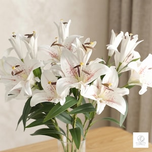 Può includere: Un bouquet di gigli bianchi artificiali con delicate macchioline rosa sui petali, stami gialli e antere marrone scuro. I fiori presentano lunghe foglie verdi appuntite su steli robusti, disposti in un vaso di vetro trasparente con logo.