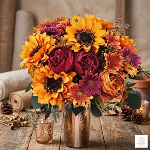 Op de afbeelding: Een levendig bloemstuk met zonnebloemen, bordeauxrode pioenrozen en oranje en paarse accentbloemen. Het boeket staat in een goudkleurige vaas, op een rustiek houten oppervlak. Een kleine kandelaar is ook zichtbaar.
