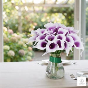 Puede incluir: Un ramo de calas artificiales con pétalos blancos que se degradan hacia centros de color púrpura intenso. Las flores están dispuestas en un florero de vidrio transparente con una cinta verde alrededor de los tallos, creando un arreglo floral.