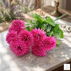 Könnte beinhalten: Ein Strauß leuchtend pinkfarbener Kunstblumen mit grünen Blättern und kleinen weißen Knospen. Die Blumen haben eine geschichtete Textur. Die Anordnung befindet sich auf einer Marmoroberfläche.