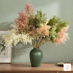 May include: A bouquet of artificial feathery foliage in shades of dusty pink, sage green, and cream, arranged in a smooth, matte dark green ceramic vase. The stems feature delicate, branching textures that create a dense, voluminous appearance.