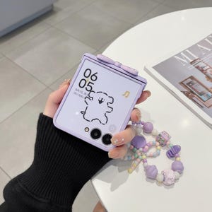 以下が含まれることがあります： 上部に小さなリボンが付いたライトパープルの折りたたみ式スマートフォンケース。画面には時刻06:05、日付、音符アイコン、クマのシンプルな黒い線画が表示されています。パステルカラーのビーズストラップが付いています。