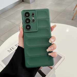 May include: A dark green smartphone case with a puffer-style design featuring horizontal padded sections. The case includes a raised rectangular camera cutout area with multiple circular lens openings arranged in a vertical and side-by-side pattern.