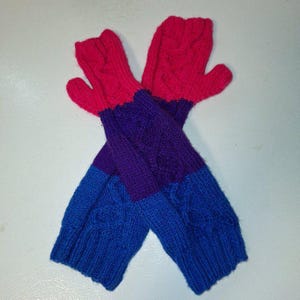 Puede incluir: Un par de mitones tejidos a mano con un diseño de bloques de color. Los mitones presentan una vibrante combinación de rojo, morado y azul. Los puños y las puntas de los dedos son azules, la sección media es morada y la parte superior es roja.