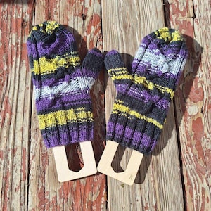 May include: A pair of knitted mittens in shades of purple, yellow, grey, and black. The mittens are displayed on wooden forms against a weathered wooden background. The mittens have a striped pattern and a ribbed cuff.