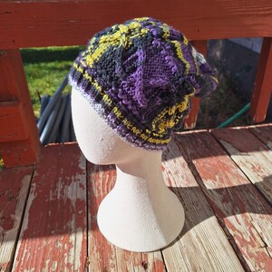 May include: A knitted beanie hat in purple, yellow, black, and grey. The hat has a textured cable knit design and is displayed on a mannequin head. The brim has a ribbed pattern.