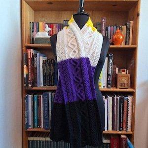 May include: A handmade, non-binary pride flag scarf. The scarf is displayed on a mannequin and features a cable knit design. The colors include yellow, white, purple, and black. The scarf is a cozy accessory.