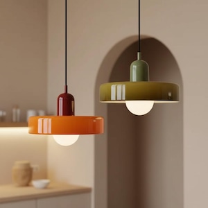 Op de afbeelding: Twee moderne hanglampen met een minimalistisch ontwerp. Eén is oranje met een bordeauxrode bovenkant, de andere is olijfgroen met een bijpassende bovenkant. Elk heeft een witte gloeilamp en een zwart snoer. Ze hangen in een keuken.