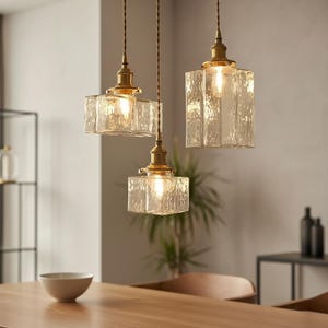May include: Three pendant lights with textured glass shades and brass-colored fixtures hang from the ceiling. The lights are rectangular with a rippled surface, and the bulbs are illuminated. The lights are suspended above a wooden table.