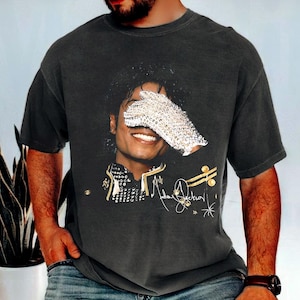 Puede incluir: Camiseta gris oscuro con un retrato de Michael Jackson, con un guante de lentejuelas cubriendo sus ojos. La camiseta incluye detalles dorados y la firma del cantante. Se ve un reloj en la muñeca.