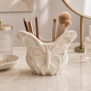 May include: A cream-colored butterfly-shaped makeup brush holder. The holder is filled with various makeup brushes and pencils. The holder is made of a stone-like material and sits on a marble surface. A mirror and perfume bottle are in the background.