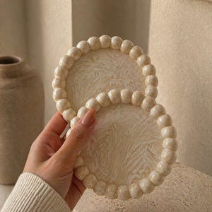 May include: Two circular coasters featuring a cream and beige marbled pattern on the flat surface, surrounded by a raised border of uniform, spherical beads in a matching marbled finish. The coasters have a smooth, glossy texture throughout.