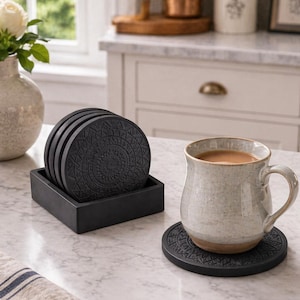 May include: A collection of black, patterned coasters in a square holder, with a mug of coffee resting on one. The coasters feature a detailed geometric design. The items are arranged on a marble surface, creating a stylish kitchen scene.